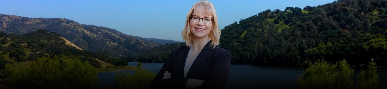 Attorney Kim Cameron-Smith standing with arms crossed in front of a scenic lake and hills